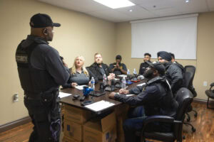 A security officer from a security guard company stands and speaks to six colleagues seated around a conference table in a meeting room., security guard companies in Toronto