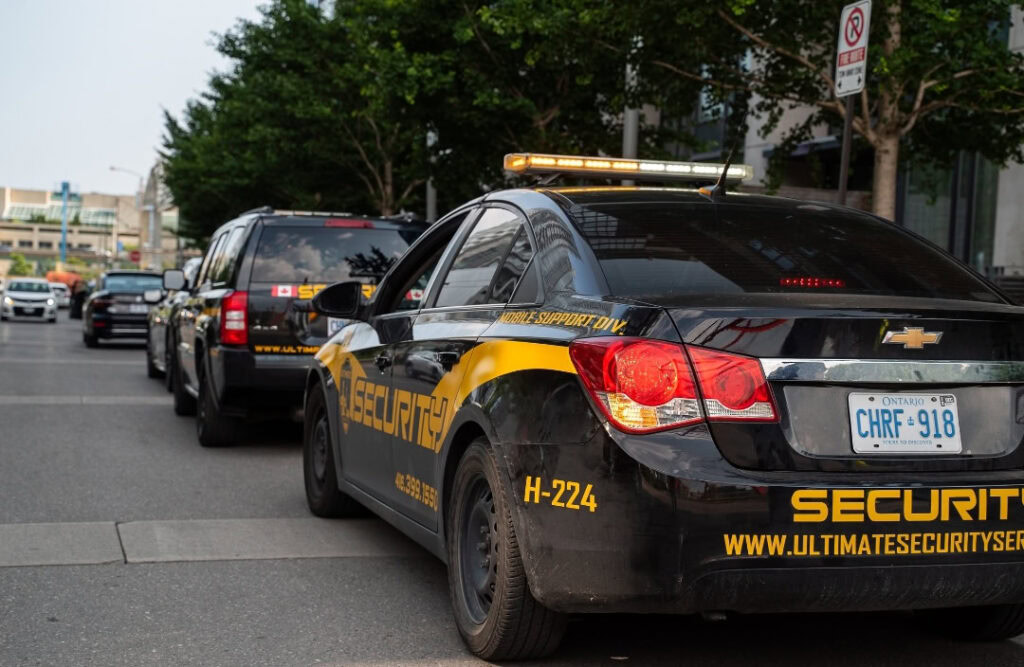 A security vehicle with flashing lights is parked on a city street, followed by another black SUV. Nearby trees and buildings are visible in the background, highlighting the presence of a Security Guard Near Me in the busy urban setting.
