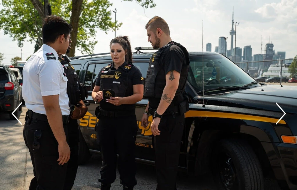 Three security officers stand and talk beside a black and yellow security vehicle in a parking lot, showcasing Security Guard Services in Toronto with city buildings in the background.
