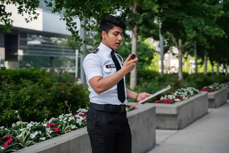 A security guard in uniform stands outdoors holding a clipboard and speaking into a handheld radio, surrounded by plants and flowers, much like a private detective in Toronto keeping a vigilant watch.