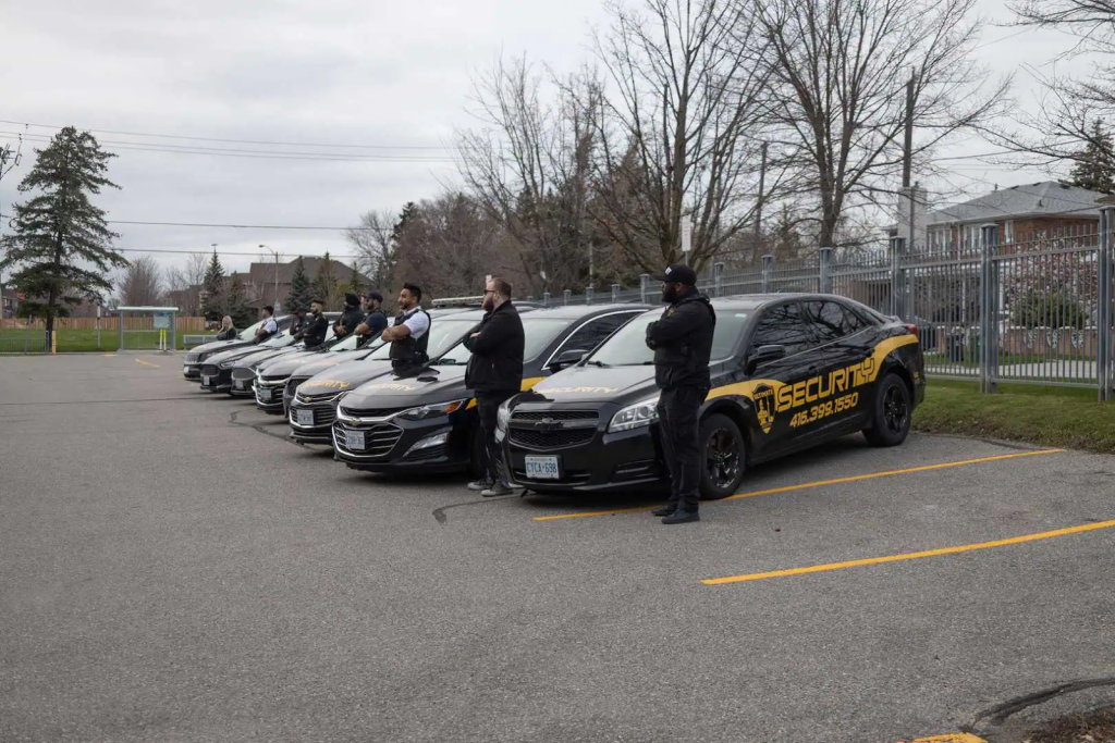 A row of security service vehicles is parked in a lot, with uniformed security guards standing beside each car on an overcast day.