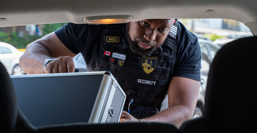 A Parking Enforcement Mississauga security guard in uniform leans into a car trunk, holding a silver briefcase, with parking lot cars visible in the background.