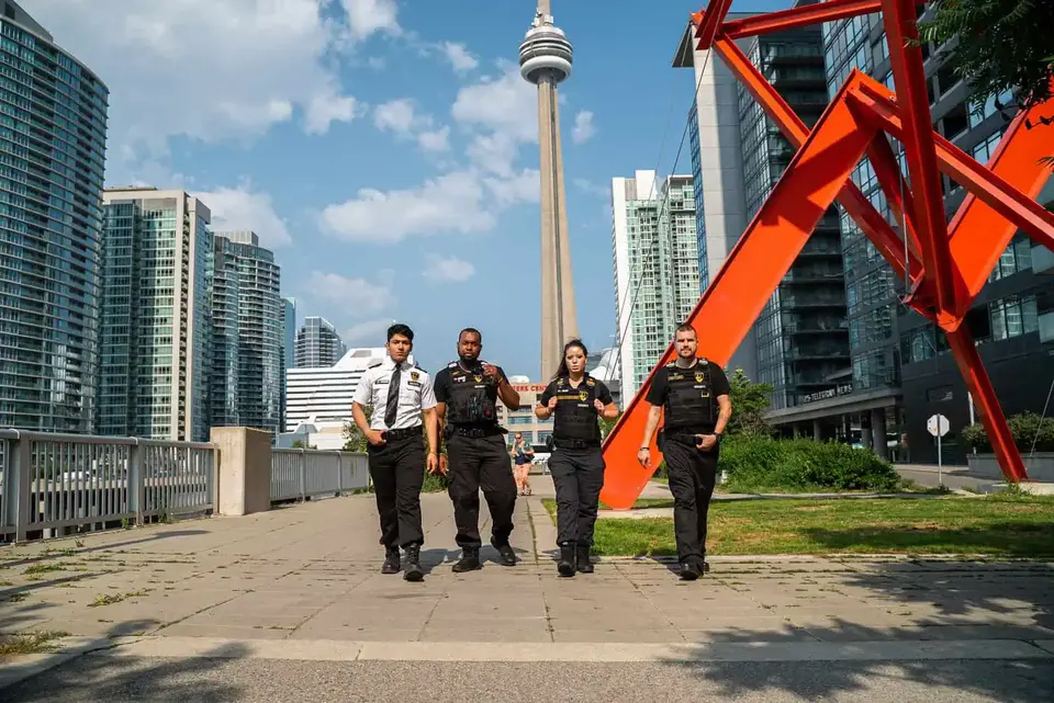 Four uniformed security officers walk on a city pathway with modern buildings and the CN Tower visible in the background, exemplifying the professionalism gained with a Security Licence in Brampton.