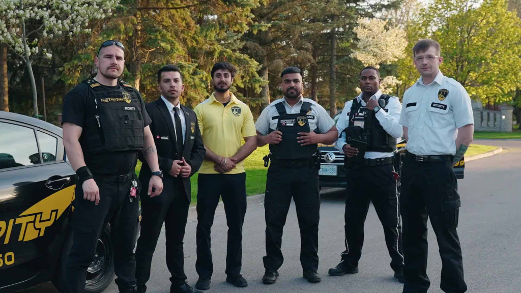 Six uniformed security guards stand in front of two parked security vehicles on a sunny day, proudly representing their Security Licence in Brampton, with trees and greenery in the background.