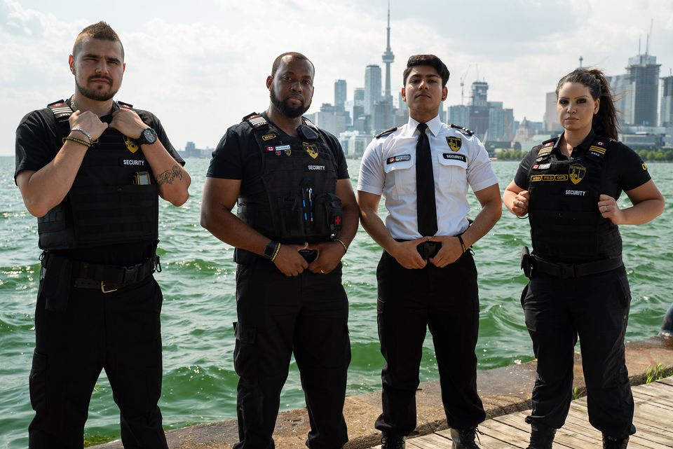 Four security officers stand side by side outdoors near a waterfront, representing Neighbourhood Watch Toronto, with a city skyline and tall buildings visible in the background.