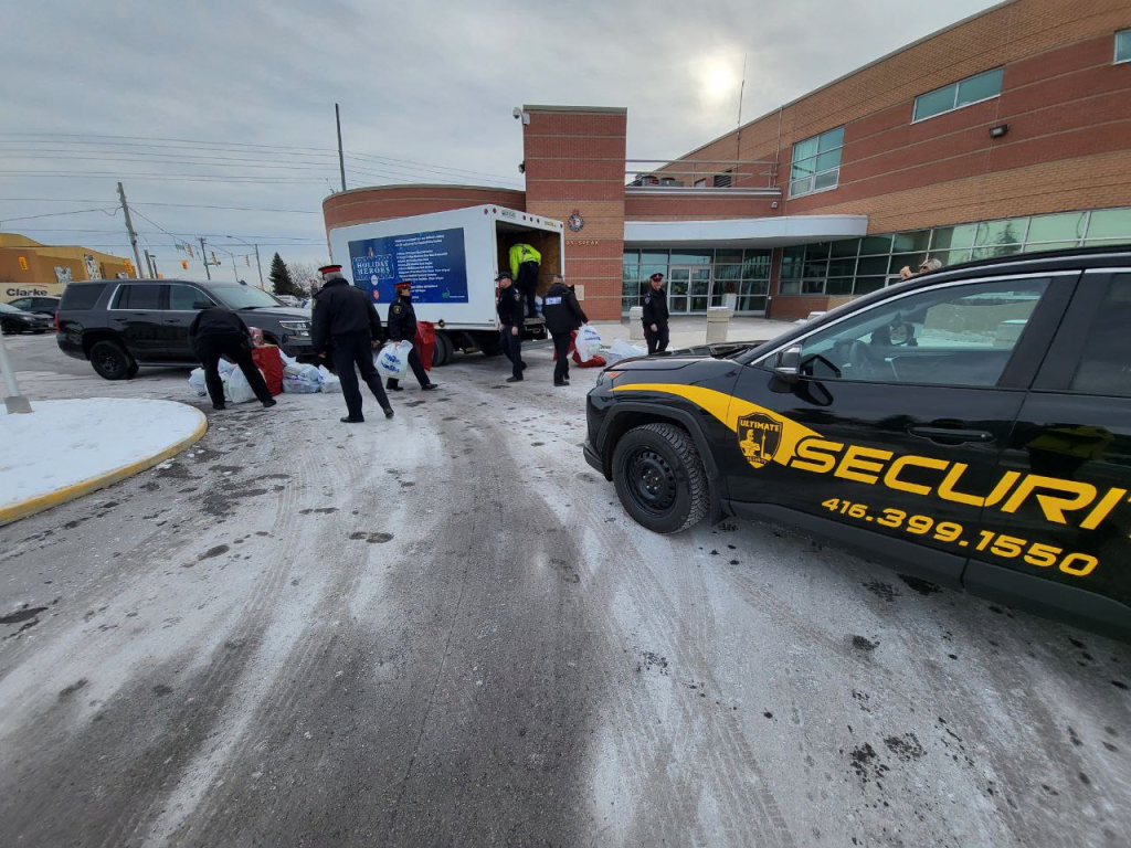 People unload items from a truck outside a building on a snowy day, with a Neighbourhood Watch Toronto security vehicle parked nearby.
