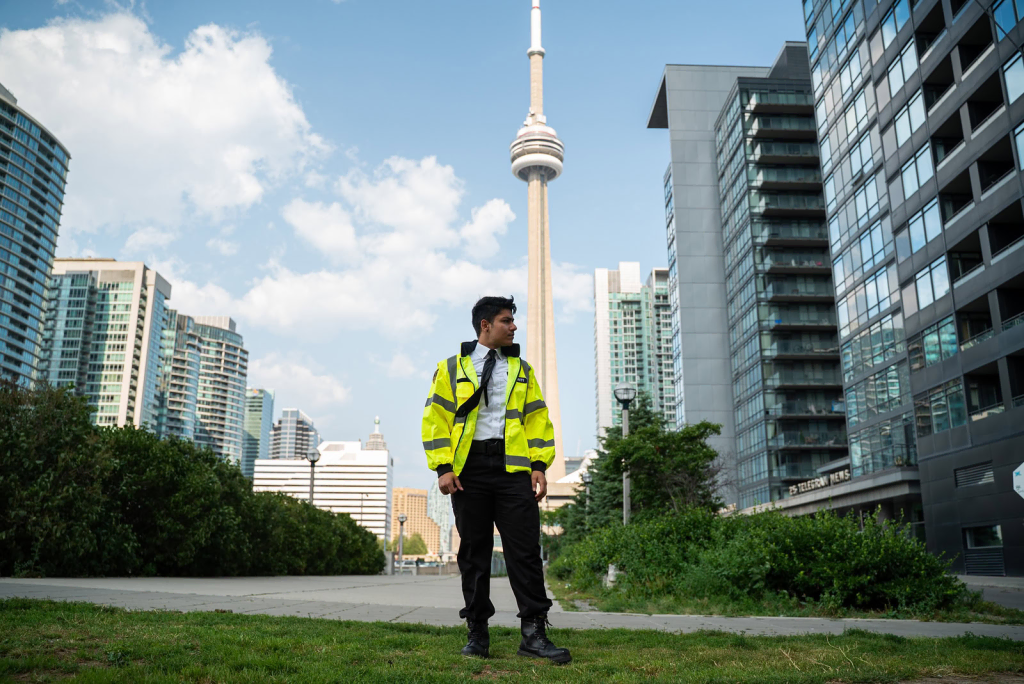A security guard in a high-visibility jacket stands on grass with tall buildings and the CN Tower in the background, representing community safety—a key element when learning how to start a neighborhood watch under a partly cloudy sky.