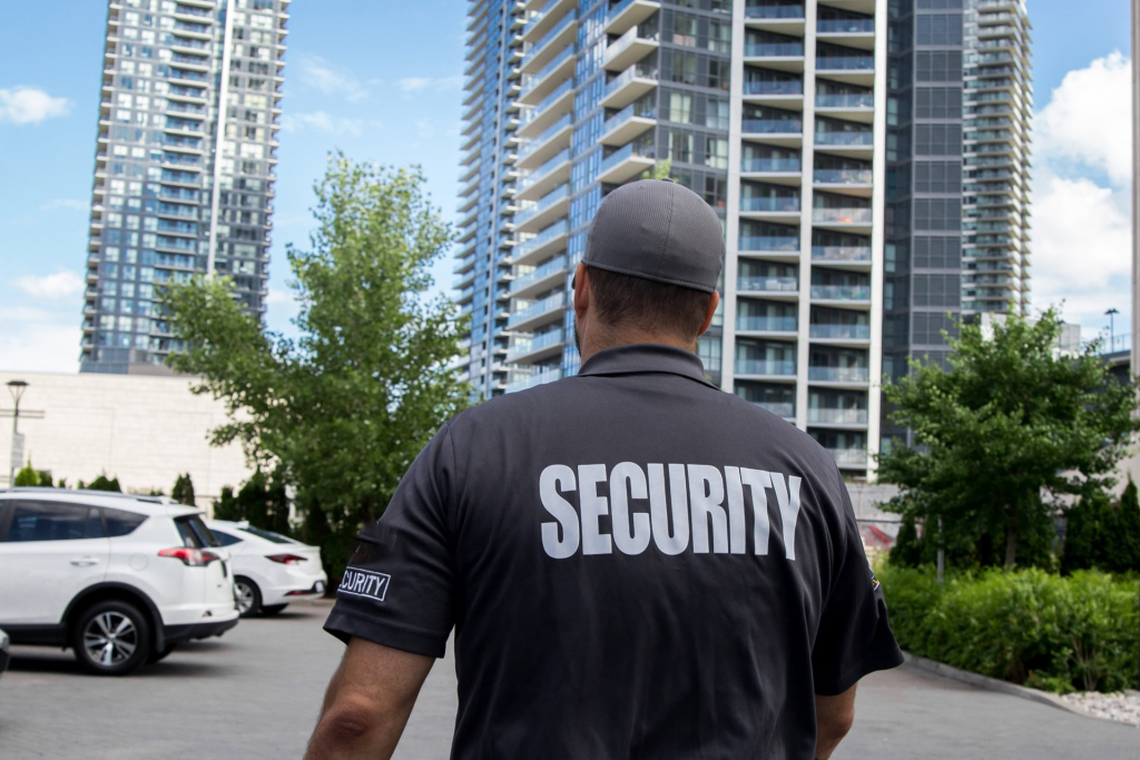 A security guard in a gray uniform stands in a parking lot near tall residential buildings on a sunny day, helping to support the Neighbourhood Crime Watch efforts.