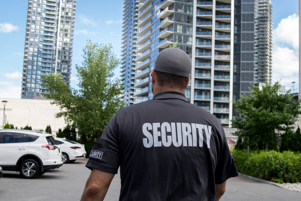 A security guard in a gray uniform stands in a parking lot near tall residential buildings on a sunny day, helping to support the Neighbourhood Crime Watch efforts.