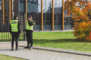 Two police officers in reflective vests stand and talk near a modern building with large windows and a tree with orange leaves, possibly discussing Neighbourhood Crime Watch efforts.
