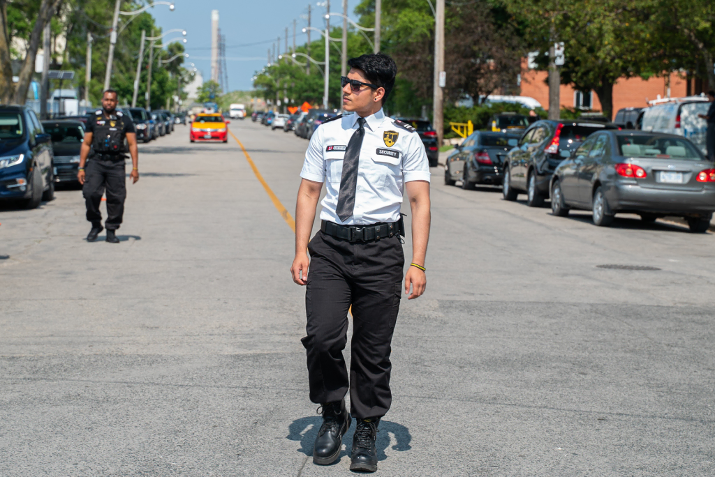 A security guard in uniform and sunglasses walks down the middle of a city street, showcasing professional Security Solutions, with parked cars and another guard visible in the background.