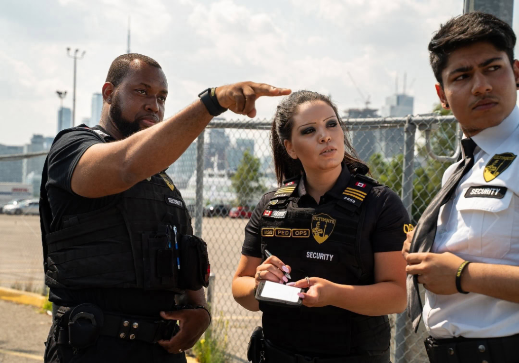 image 12 Three security personnel from Affordable Security Solutions stand outdoors; one points into the distance, another writes on a notepad, and the third looks to the side. A city skyline and fence can be seen in the background.