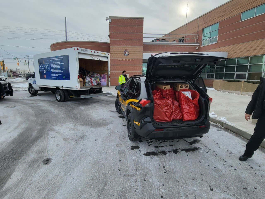 image 13 A police SUV and a delivery truck, both marked with Affordable Security Solutions, are parked outside a brick building on a snowy street, their trunks open and filled with large red bags and boxes.