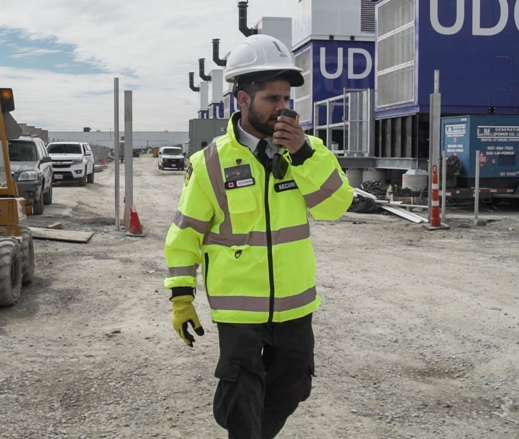 A security guard in a high-visibility jacket and hard hat speaks into a handheld radio while walking on a construction site, representing a trusted Security Service in Toronto.