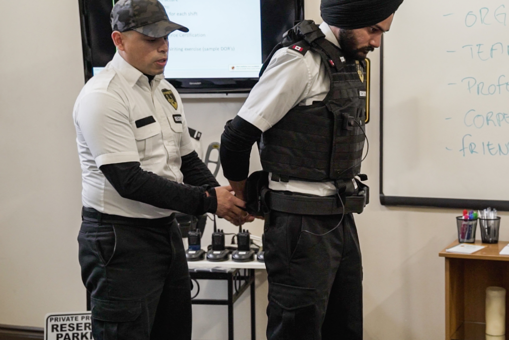 Two security officers in uniform, practicing basic security guard training in Ontario, stand in a room with a whiteboard and communication devices visible in the background.