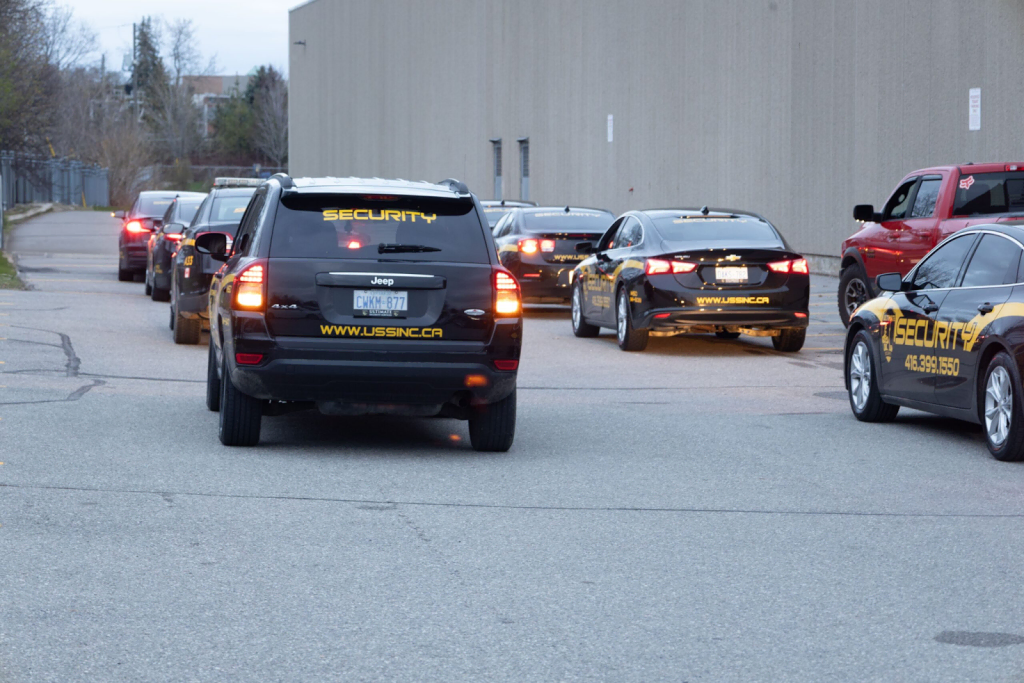 A line of security vehicles with "SECURITY" and company branding, operated by professionals with basic security guard training in Ontario, are parked and driving along a paved street next to a large beige building.