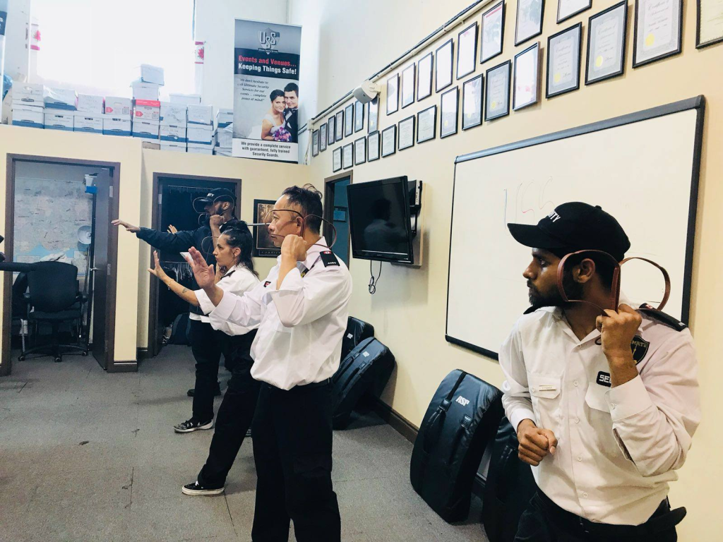 Four security guards from a security company near me practice defensive stances and radio use in a training room with certificates on the wall and storage boxes stacked above shelves.