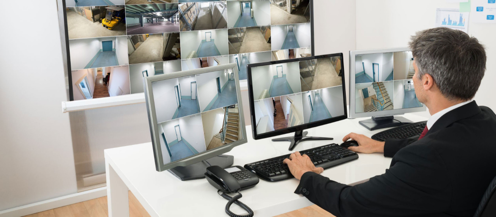 image 21 A man in a suit monitors multiple security camera feeds on three computer screens and a large wall display in a surveillance control room at a leading Security Company in Vaughan.