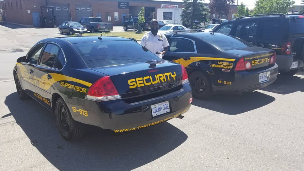 image 5 Two security cars marked "SECURITY" and "SUPERVISOR" are parked side by side in a lot, showcasing Parking Enforcement in Mississauga. A uniformed security officer stands between them, with buildings visible in the background.